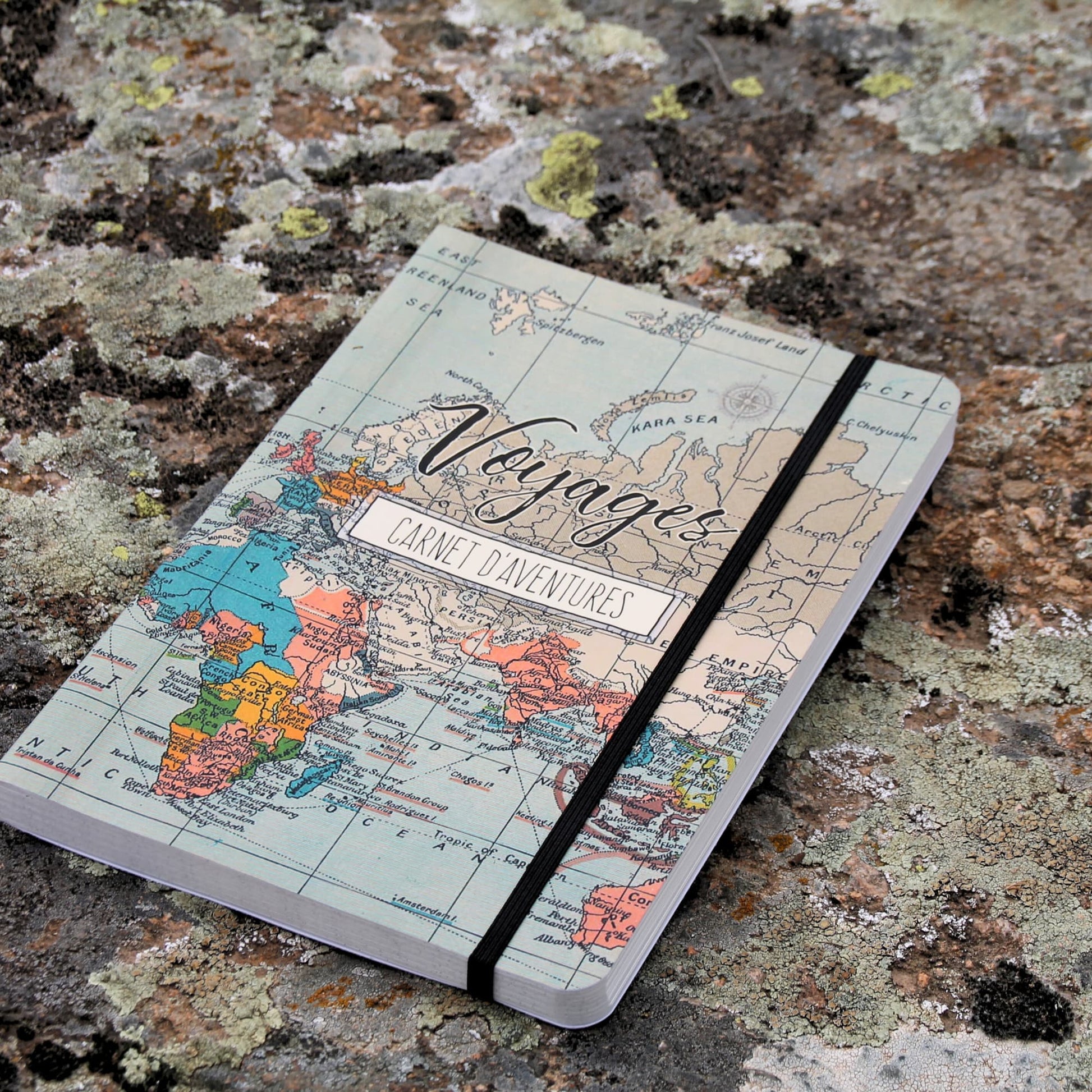 Grand carnet de voyage format A5 fermé, avec une couverture illustrée de motifs de voyage et de mappemonde, maintenu par un élastique.