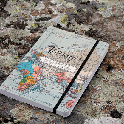 Grand carnet de voyage format A5 fermé, avec une couverture illustrée de motifs de voyage et de mappemonde, maintenu par un élastique.
