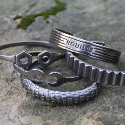 Ambiance avec 5 bracelets joncs en acier inoxydable argenté, représentant des écrous, des clés plates, des engrenages et un moteur, de la marque Rouille.