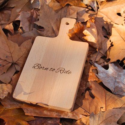 Petite planche à découper en bois gravé Born to Ride, posée dans des feuilles pour une ambiance automnale.