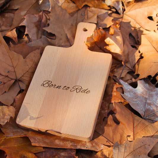 Petite planche à découper en bois gravé Born to Ride, posée dans des feuilles pour une ambiance automnale.