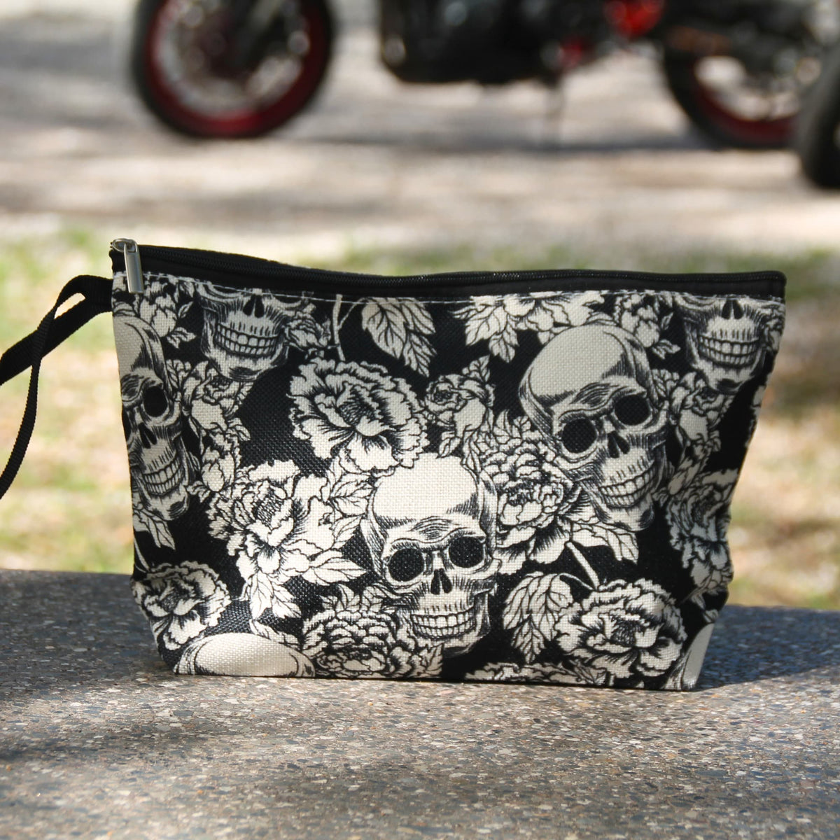 Pochette noire à crânes blancs et roses présentée devant une moto, idée cadeau pour motarde au style rock.