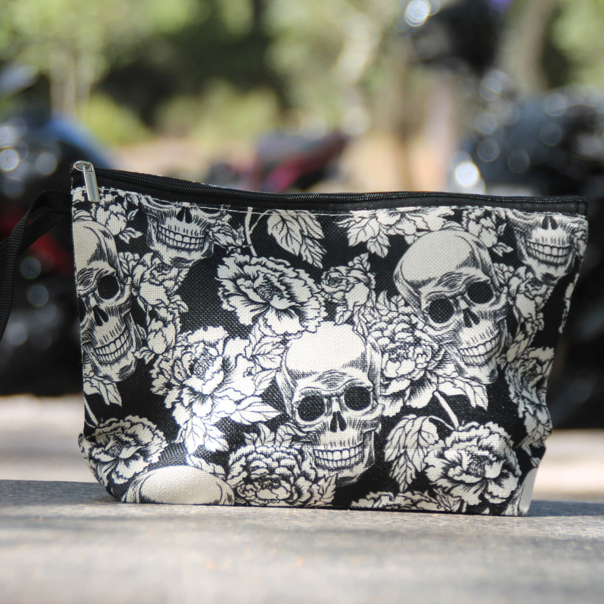 Pochette noire à crânes blancs et roses présentée devant une moto, idée cadeau pour motarde au style rock.