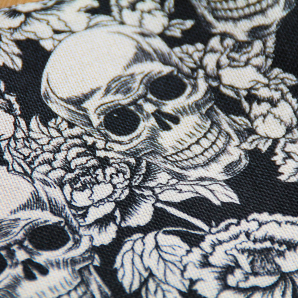  Gros plan sur le tissu texturé effet lin et les motifs de crânes blancs, roses et feuillages de la pochette noire.