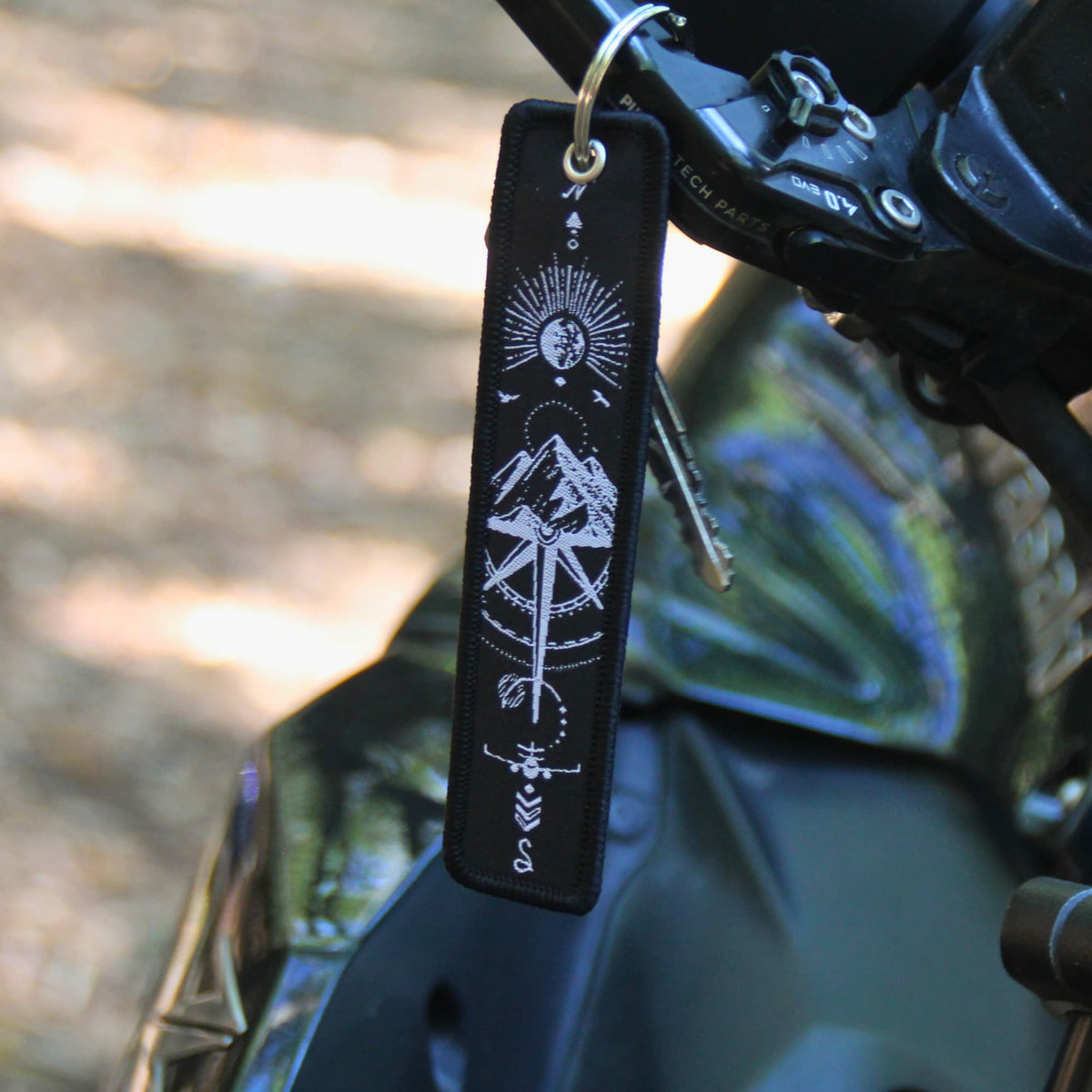 Porte clé moto en tissu noir brodé de symboles evoquant le voyage et l'évasion (rose des vents, soleil, montagne, oiseaux...)